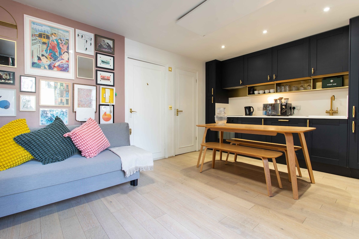 A modern living area showcases a light gray sofa adorned with colorful decorative cushions. A wooden dining table with benches is positioned nearby, complemented by stylish kitchen cabinetry in navy blue. Various framed artworks add visual interest to the wall.