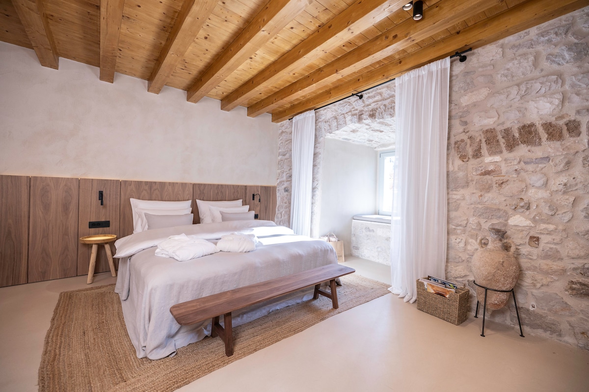 A spacious bedroom features light-colored walls and exposed wooden beams. A large, cozy bed is adorned with soft linens, and a bench sits at the foot. Natural light enters through a window draped with sheer curtains, creating a calm and inviting atmosphere.