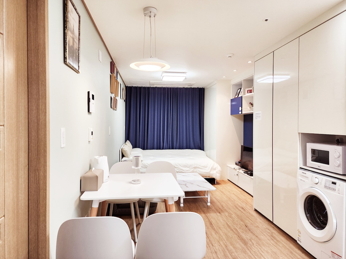 A modern interior showcases a compact living space with light wood flooring. A spacious bed is positioned against a blue curtain backdrop. A white dining table with four chairs is to the front, while kitchen and laundry amenities are integrated into the cabinetry.