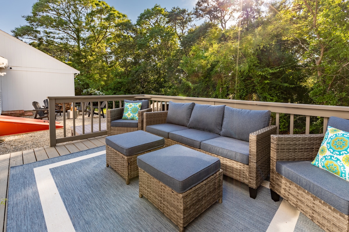 Outdoor lounge seating is arranged on a patio with a neutral rug. Cushions adorned with colorful patterns add a touch of comfort. Lush greenery surrounds the area, creating a serene atmosphere, while sunlight filters through the trees, enhancing the inviting setting.