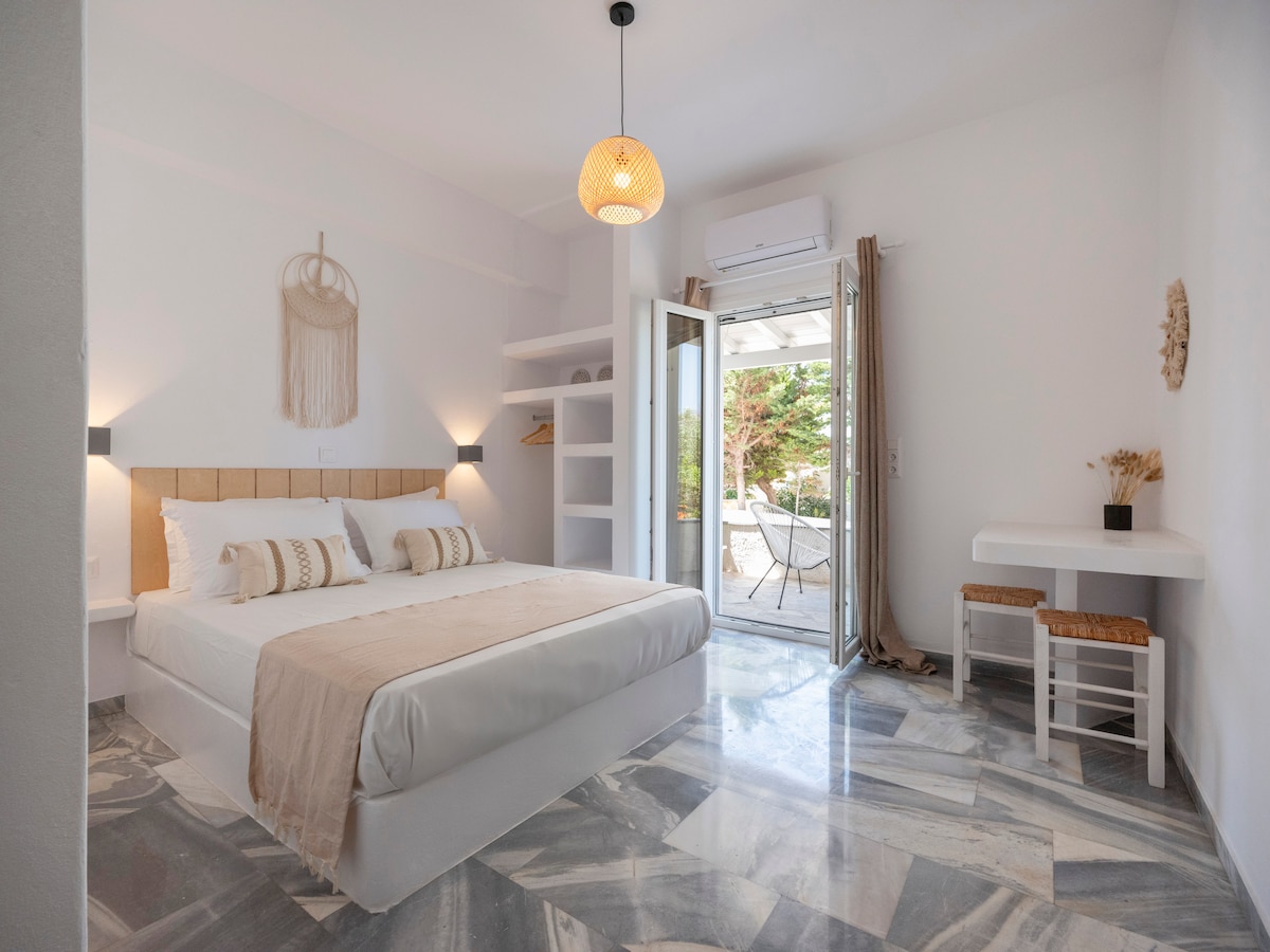 A light-filled bedroom features a spacious bed with neutral-toned bedding and decorative pillows. Smooth marble flooring complements the minimalist design. A small table and chairs are positioned near glass doors, which open to a terrace with outdoor seating and a garden view.