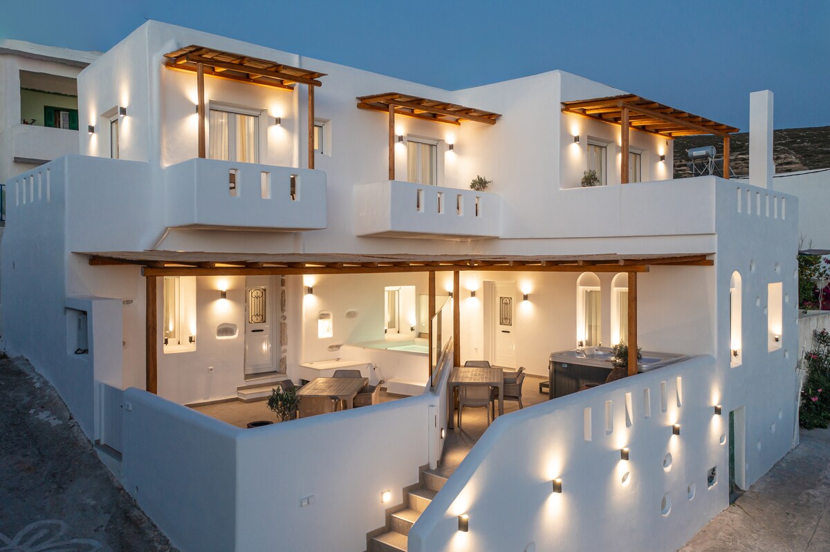 A modern building with a minimalist design features multiple levels and a spacious terrace. Subtle lighting accentuates the exterior, creating a welcoming ambiance. A hot tub is visible on the terrace, surrounded by outdoor seating and greenery, inviting relaxation and enjoyment.