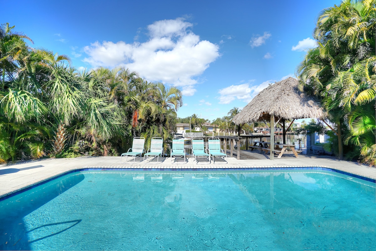 A sunlit pool area features a glistening swimming pool surrounded by lush greenery. Lounge chairs are positioned around the pool, and a thatched Tiki hut offers shade. The sky above is clear, with a few clouds adding texture to the scene.