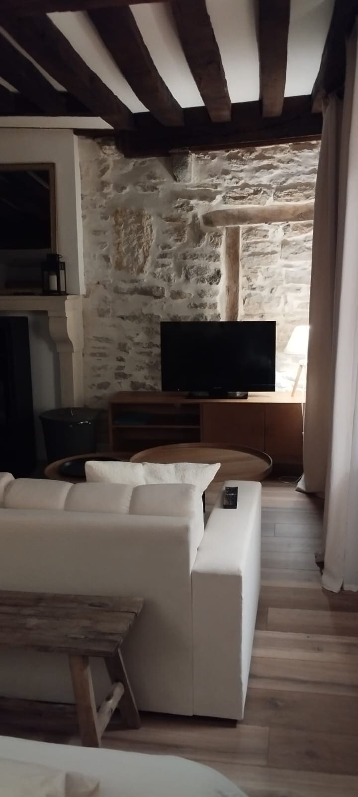 A cozy living area features a white sofa positioned next to a wooden coffee table. A television is mounted on a natural wood console against a textured stone wall, contrasting with the wooden beams overhead.