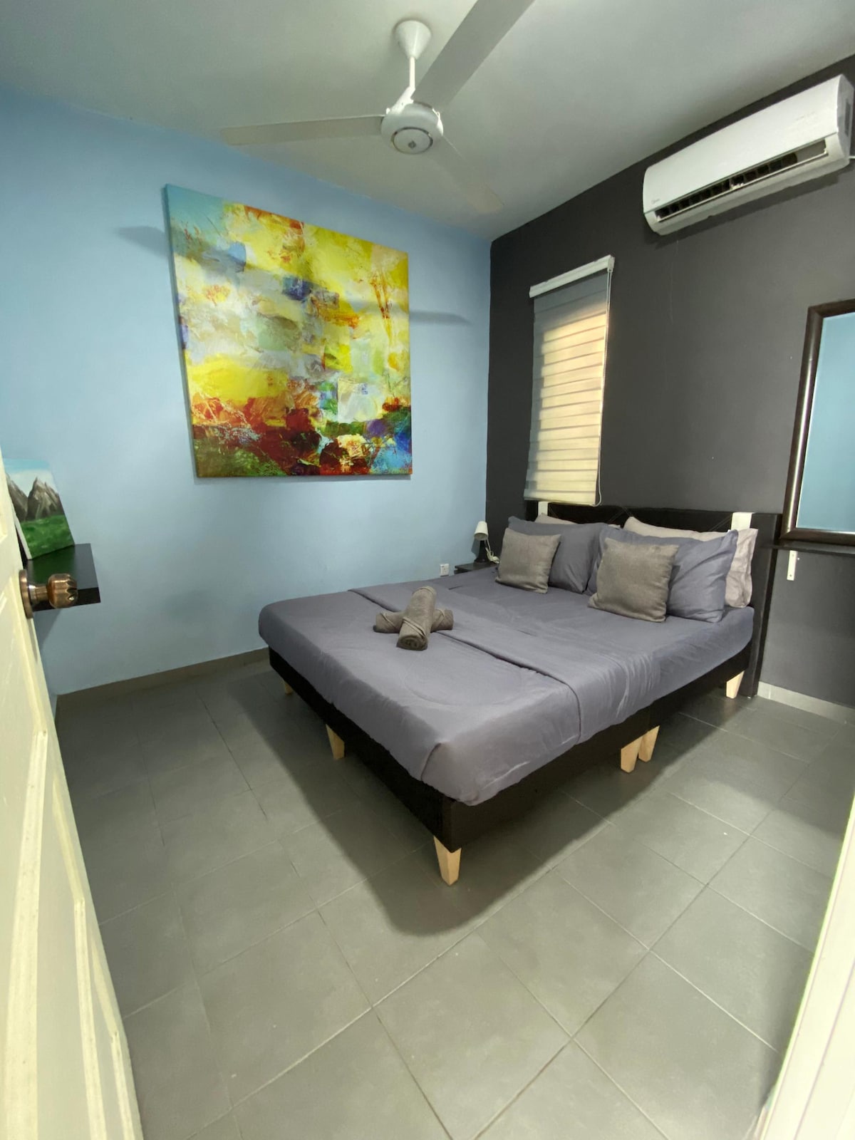 A cozy bedroom features a spacious bed with gray linens and decorative pillows. An abstract painting in warm colors adorns the wall above the bed. Natural light filters through the partially covered window, highlighting the cool blue and dark gray walls.