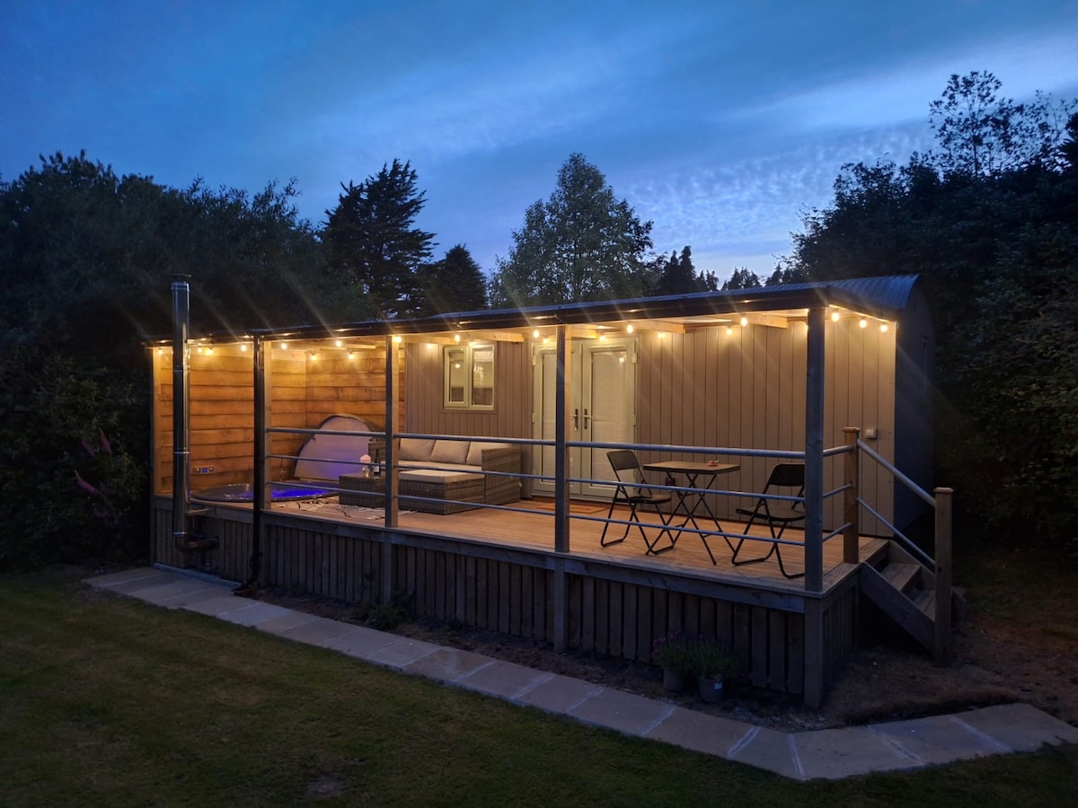 A wooden deck is illuminated by string lights, featuring a comfortable seating area with a sofa and two chairs. A table is positioned nearby for outdoor dining. Surrounding greenery enhances the secluded feel of the space, with a door leading into the cabin.