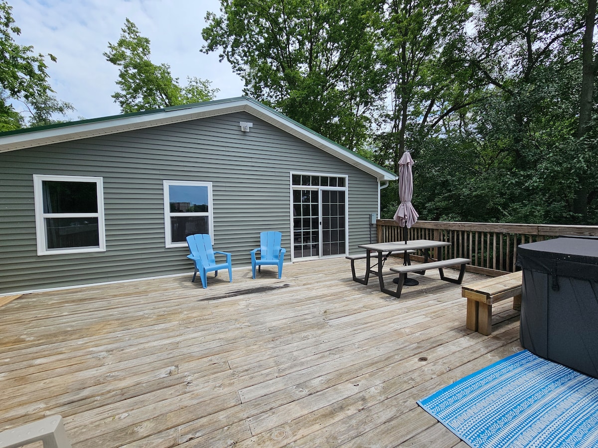 The spacious back deck features seating with two blue adirondack chairs and a picnic table. A hot tub is positioned near the railing, providing views of the surrounding trees. A large umbrella offers shade, while a patterned rug adds color to the wooden surface.