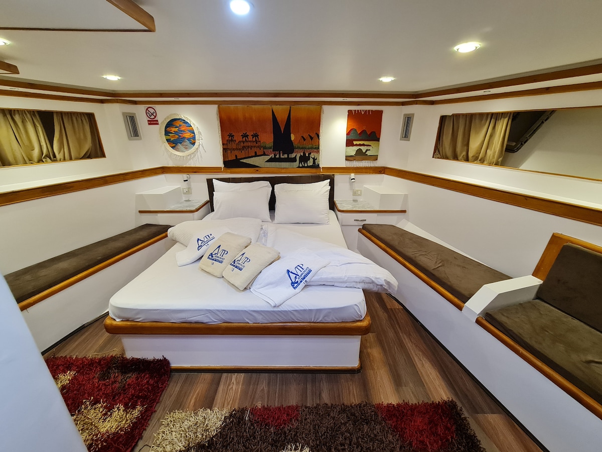 A cabin features a spacious bed with clean white linens and decorative pillows. Two upholstered seating areas are positioned against the walls. Artwork depicting boats and a sunset adorns the walls. Soft lighting creates a calm ambiance, complemented by plush rugs on the wooden floor.