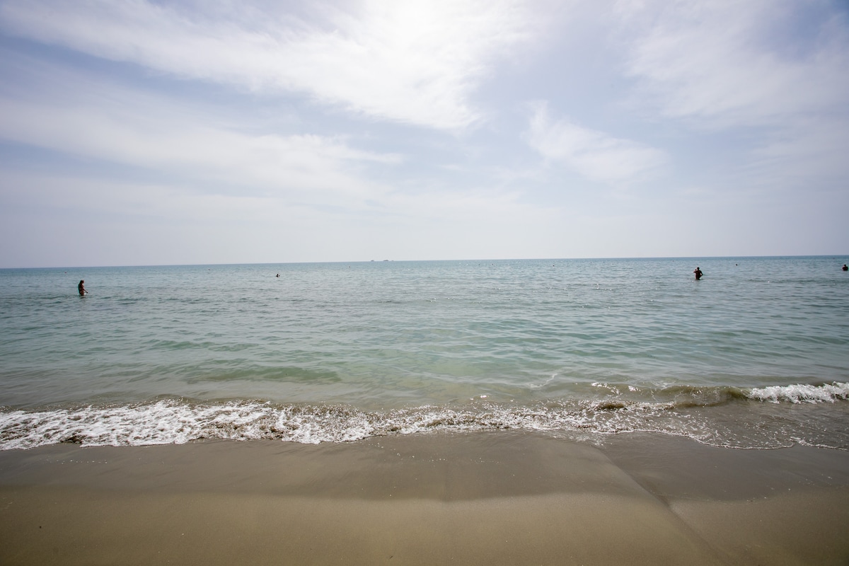 The serene beach scene captures gentle waves lapping against a sandy shore, with a calm sea stretching to the horizon. A few figures can be seen in the water, enjoying the tranquil atmosphere under a bright sky with scattered clouds.