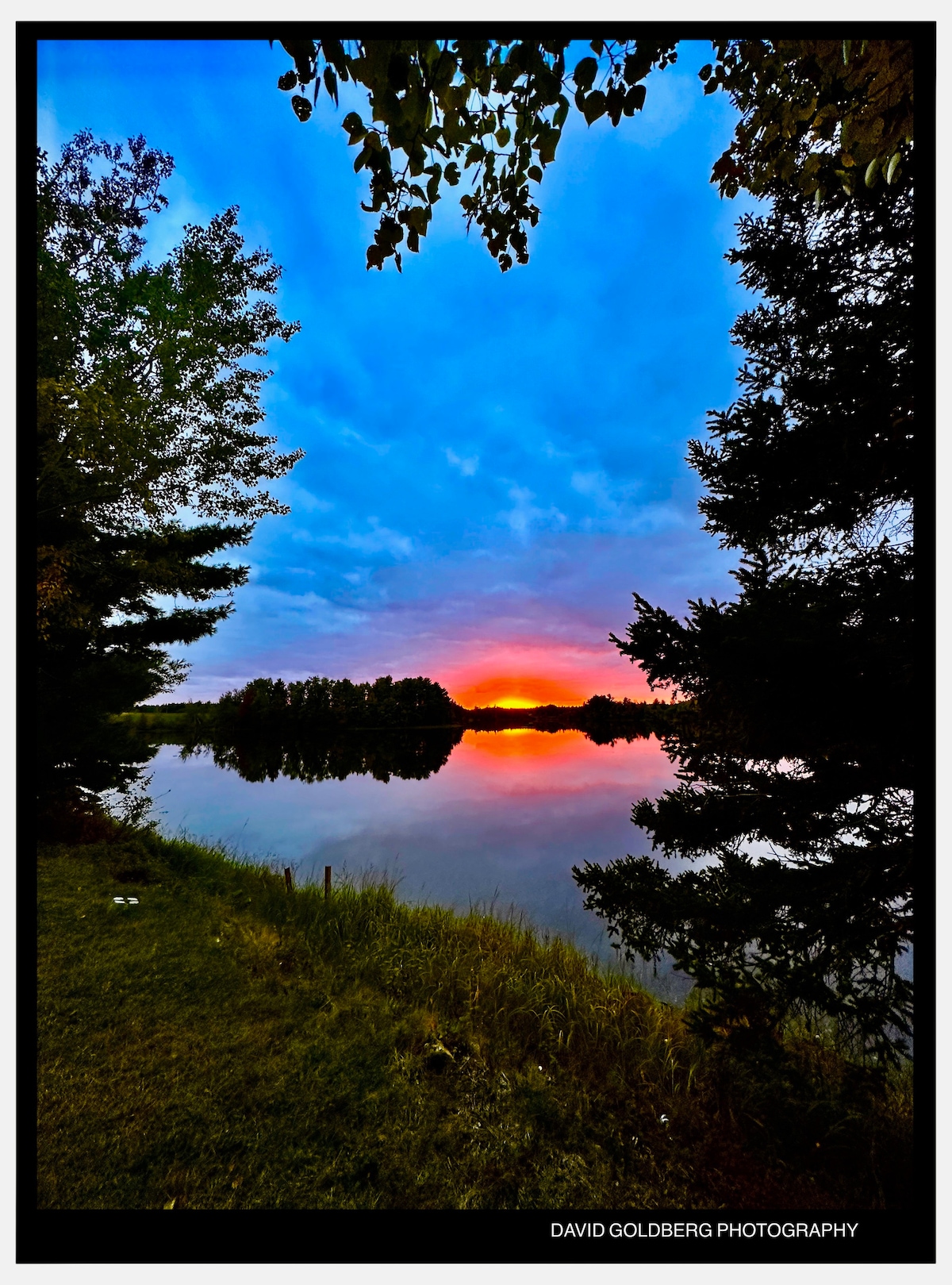 A serene view of the water reflects a colorful sunset, framed by trees on either side. The sky is filled with varying shades of blue and orange, creating a peaceful atmosphere by the water's edge.