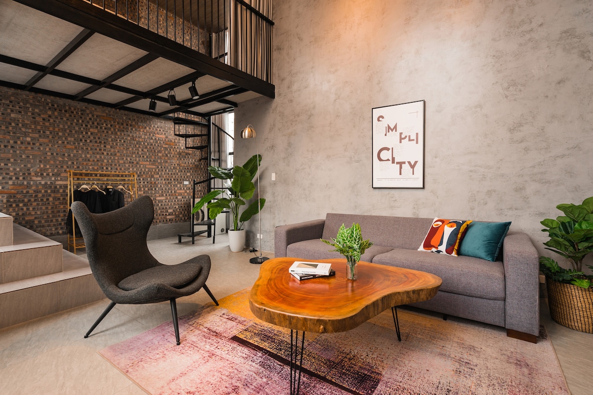 A modern living area features a charcoal gray sofa and an artistic wooden coffee table. A textured wall in warm tones is complemented by green plants and a decorative wall art piece. Soft lighting accentuates the open layout, with a spiral staircase visible in the background.