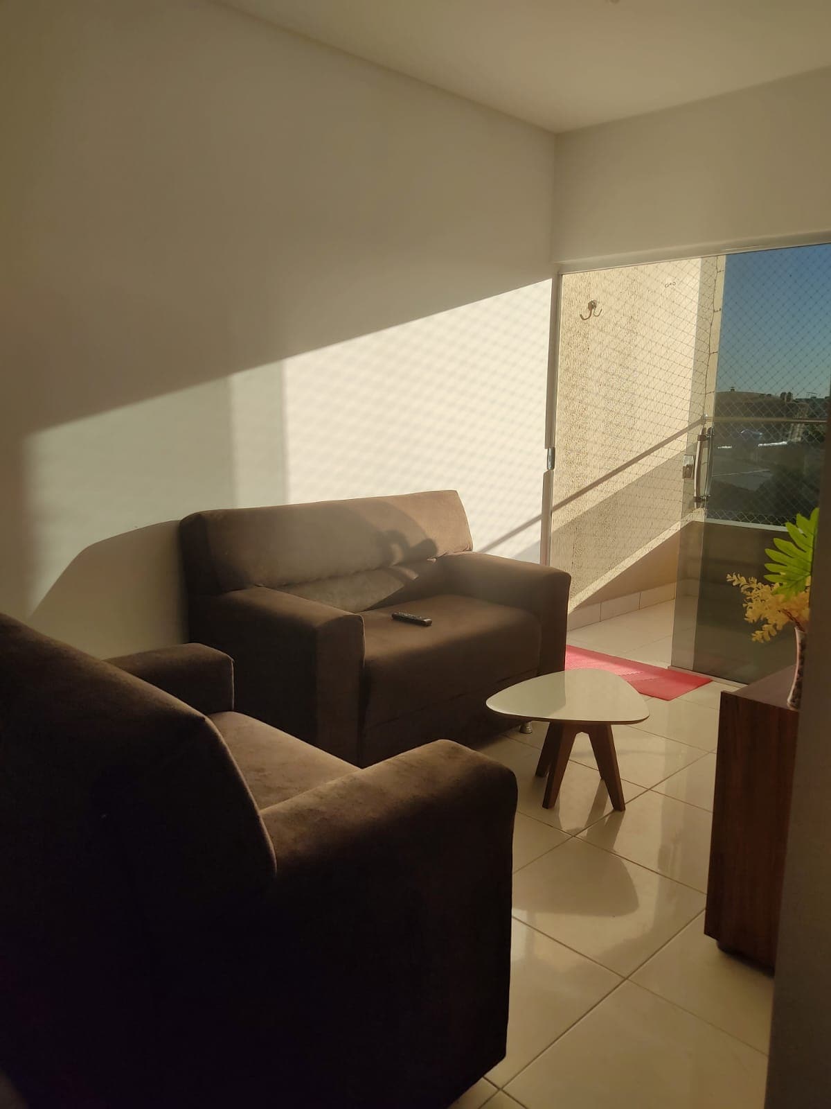 A cozy seating area is showcased with two brown armchairs positioned beside a small wooden table. Natural light filters through the nearby doorway, casting soft shadows on the tiled floor. A decorative plant adds a touch of greenery, enhancing the space's tranquil atmosphere.