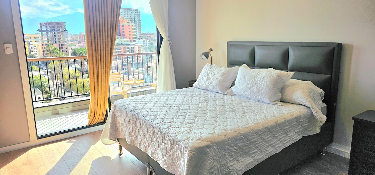 A bright bedroom features a spacious bed with crisp white bedding. Large windows allow natural light to fill the space and offer views of the cityscape. A small balcony with outdoor seating can be seen through the open curtains.