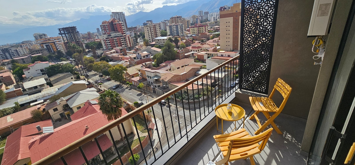 A balcony offers a view of the urban landscape, featuring a mix of residential buildings and vibrant city life. Two yellow wooden chairs sit beside a small round table, inviting relaxation while overlooking the scenic surroundings.