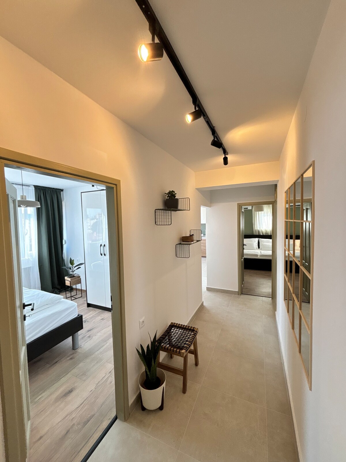 A well-lit hallway features a combination of light and neutral tones. An inviting seating area is positioned against one wall, complemented by a small plant. Decorative shelving holds minimalistic items, while a large mirror enhances the sense of space, leading to a glimpse of the spacious bedroom.
