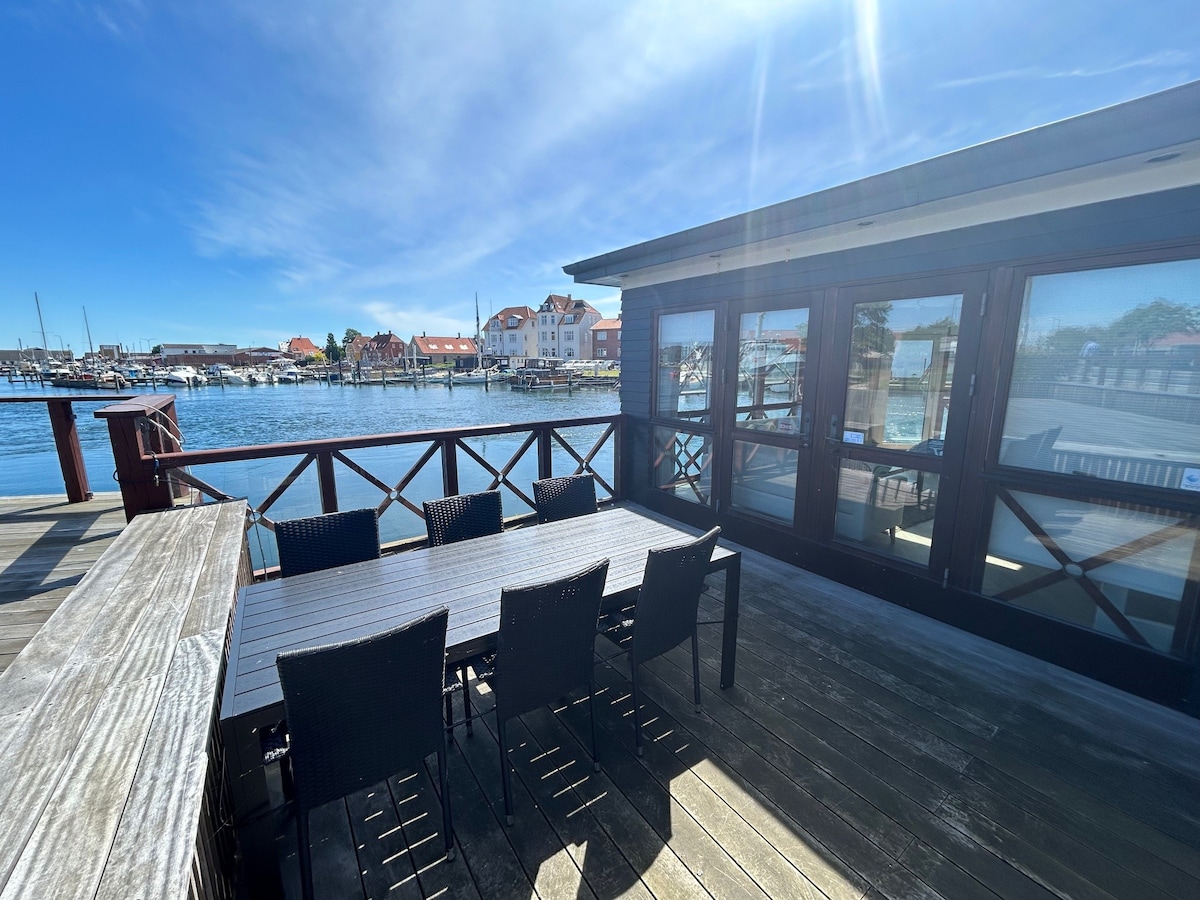 A spacious outdoor terrace is featured, equipped with a dining table and chairs facing the water. Large windows provide views of the marina and surrounding buildings, while sunlight reflects off the water's surface, creating a bright and inviting environment.