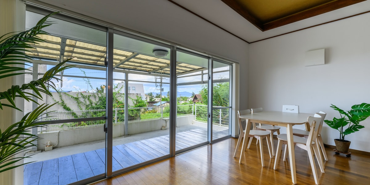 A spacious room features large sliding glass doors that provide access to a terrace with wooden flooring. A simple dining table with chairs is positioned nearby. Greenery from potted plants adds a touch of nature, while natural light fills the space.
