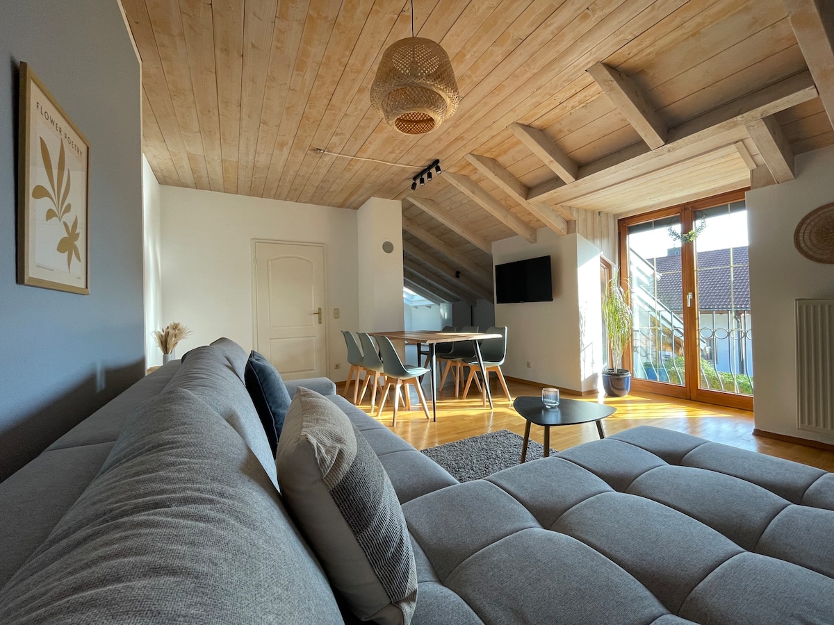 A spacious living area features a large gray sofa with decorative pillows, complemented by wooden ceiling beams. Natural light filters through the glass doors, which lead to a balcony. A dining table with chairs is positioned nearby, offering a cozy environment for gatherings.