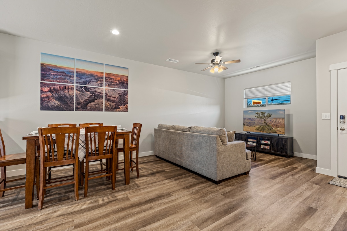 An open living area features a comfortable couch and a dining table with four wooden chairs. A television unit is situated opposite the seating area. Wall art depicting a landscape is displayed above the table, and natural light filters through a window with blinds.