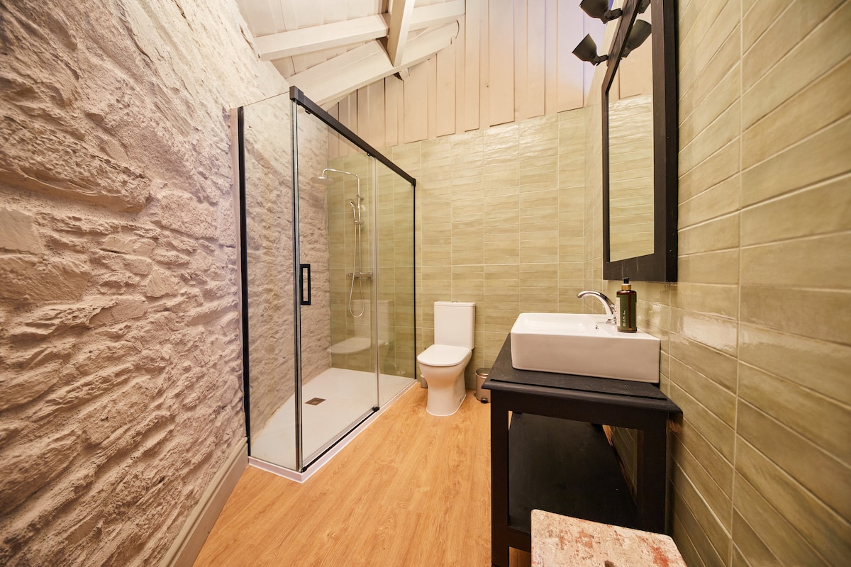 A modern bathroom features a spacious walk-in shower enclosed by clear glass panels. A white toilet is situated beside a sleek sink with a minimalist design. The walls are adorned with subtle green tiles, while the wooden floor provides a warm contrast to the contemporary fixtures.