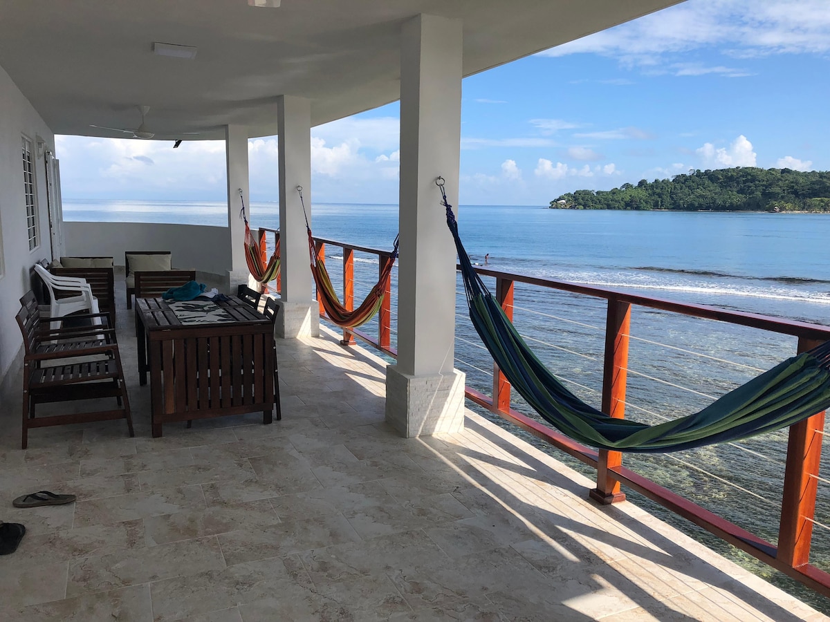 A spacious terrace overlooks the serene sea, featuring comfortable seating and colorful hammocks gently swaying. The panoramic view includes distant green hills, inviting relaxation. The flooring is elegantly tiled, contributing to a modern yet comfortable outdoor space for guests.