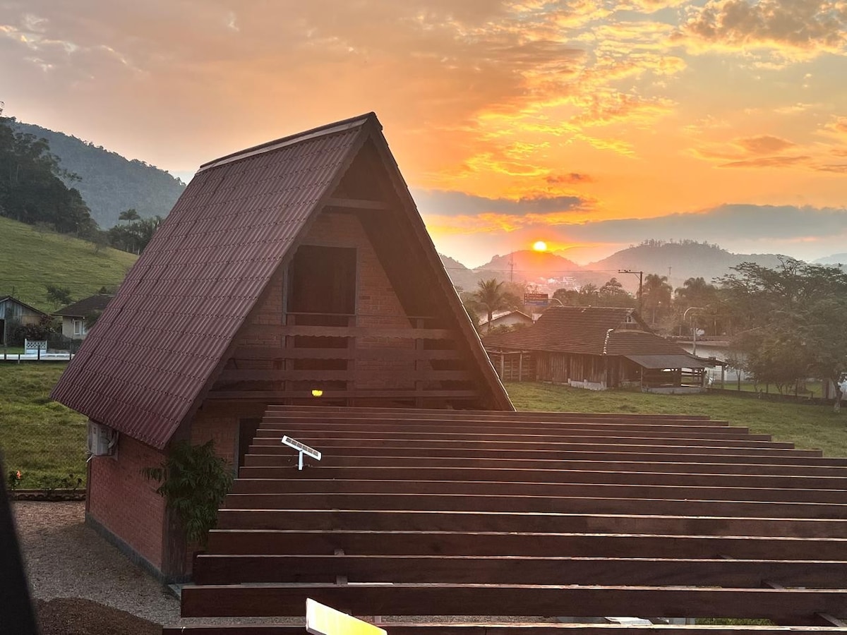 An A-frame chalet is viewed against a vibrant sunset, highlighting the roof's sloped design and wooden beams. The surrounding landscape features gentle hills and a lush green area, contributing to a serene atmosphere. A structured outdoor space is visible, providing ample room for relaxation.