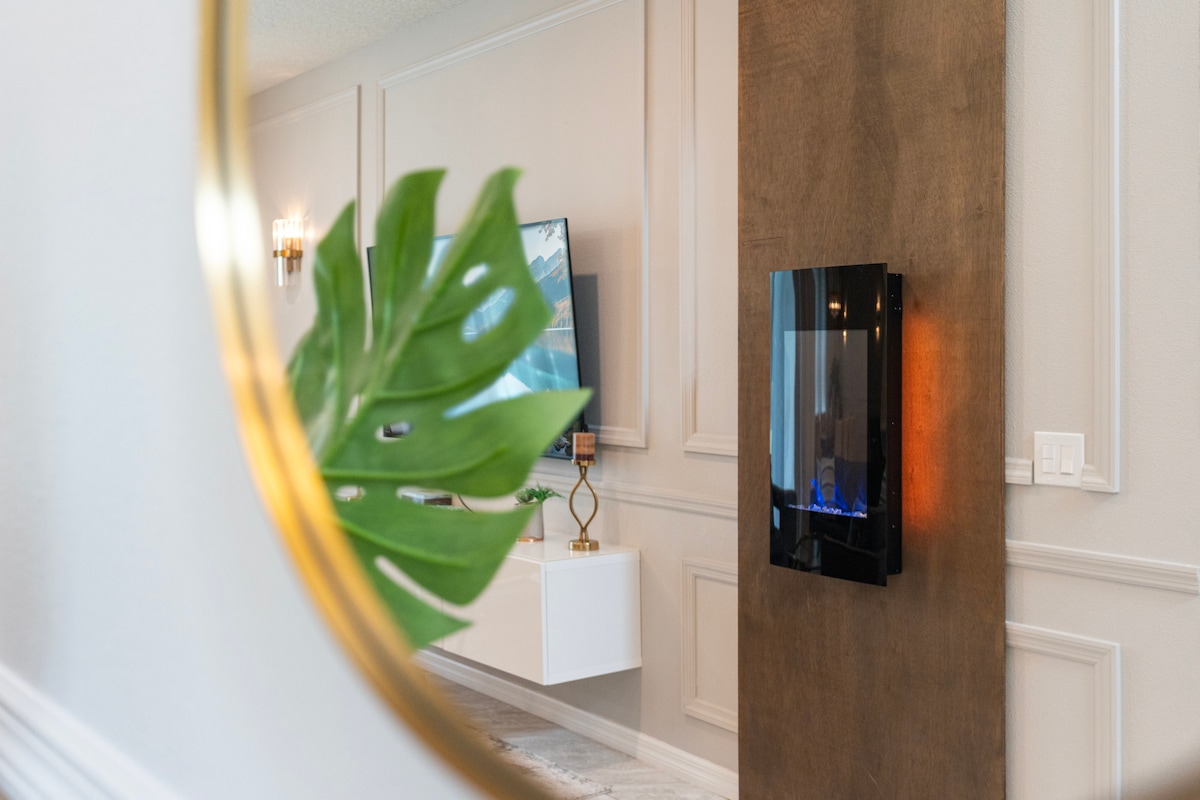 A modern living space is depicted with a sleek electric fireplace mounted on a feature wall. Decorative elements are present, including a leaf from a plant seen in the foreground, and a stylish wall-mounted television is positioned in the background.