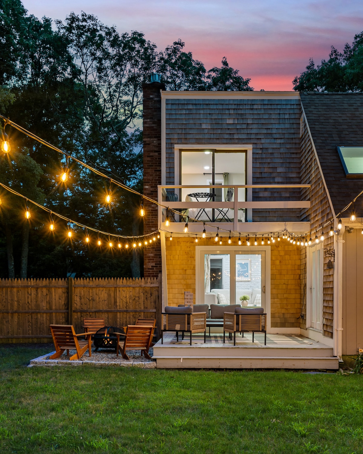 The exterior of a modern home is highlighted by warm exterior lighting, creating a welcoming ambiance. A spacious patio area hosts comfortable seating, while strung bistro lights above enhance the outdoor space. Lush green grass surrounds the patio and leads to a fenced yard.
