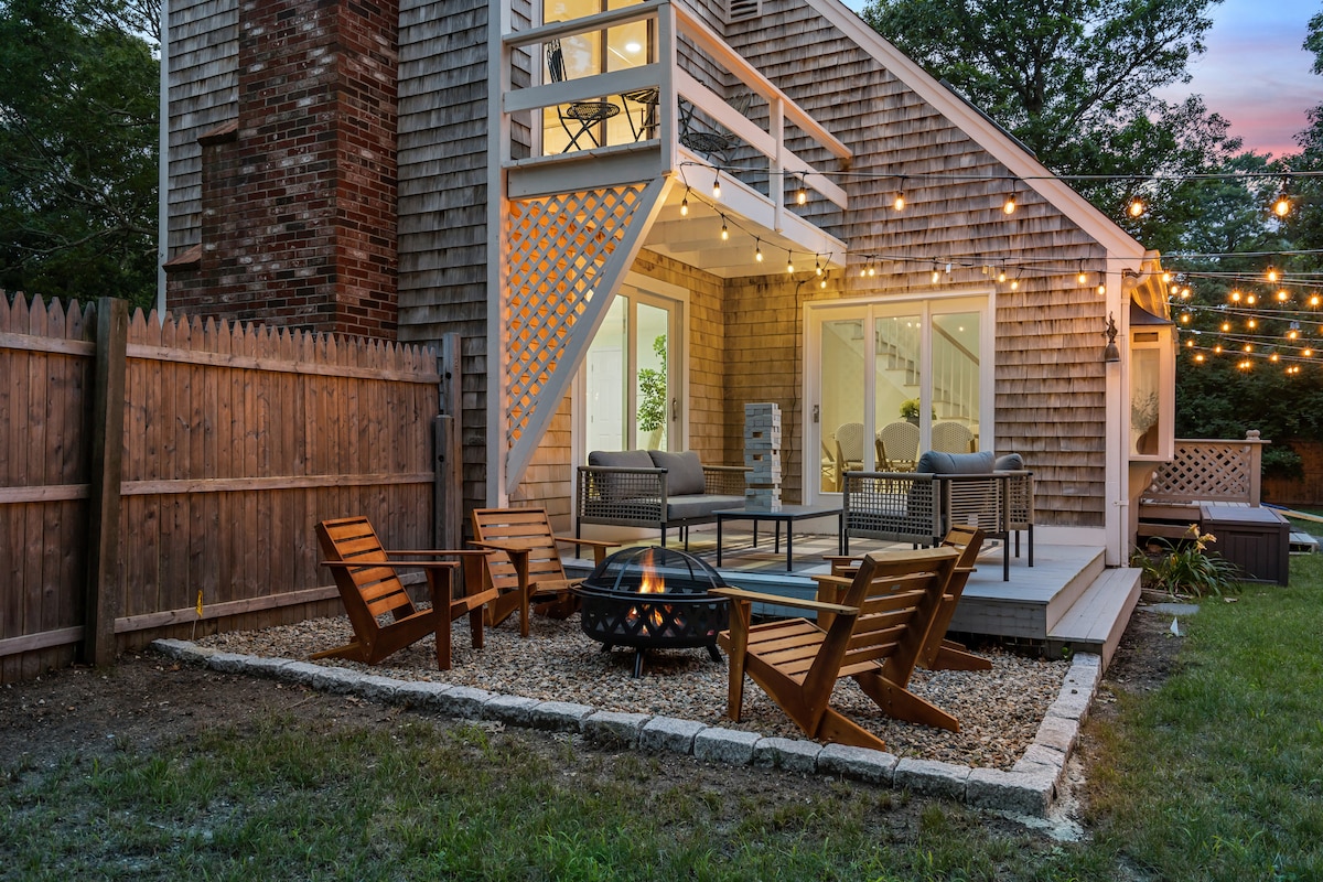 A cozy outdoor seating area is highlighted by four wooden chairs arranged around a gravel fire pit. The exterior of the house features large sliding glass doors that connect to a welcoming deck, adorned with bistro lights for a relaxed ambiance.
