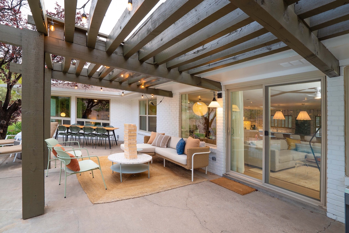 An outdoor living space is defined by a spacious tiled patio covered by a wooden pergola. A comfortable seating area features a circular coffee table surrounded by cushioned chairs, while a dining set is positioned nearby. Large sliding glass doors provide access to the interior.