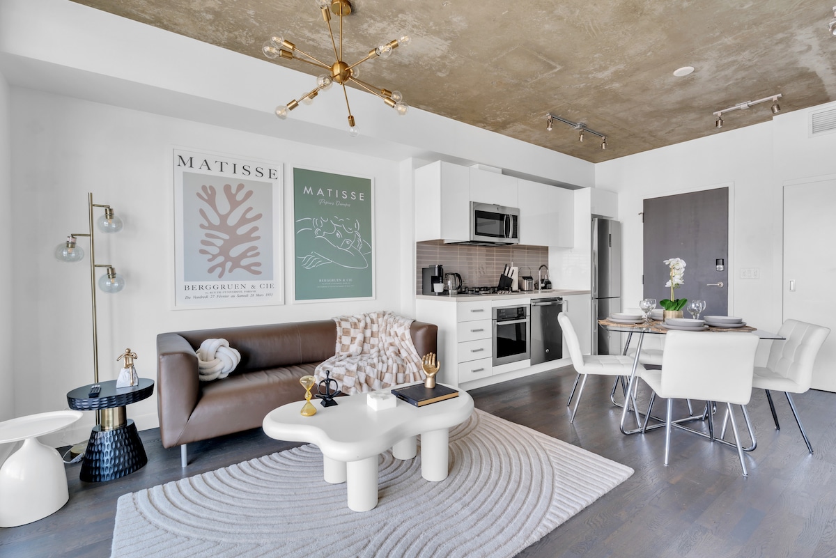 A modern living space is showcased, featuring a stylish brown sofa and a unique white coffee table. Artwork by Matisse adorns the walls. A contemporary dining area with white chairs and a sleek table is visible. The kitchen area, equipped with modern appliances, is positioned at the back.