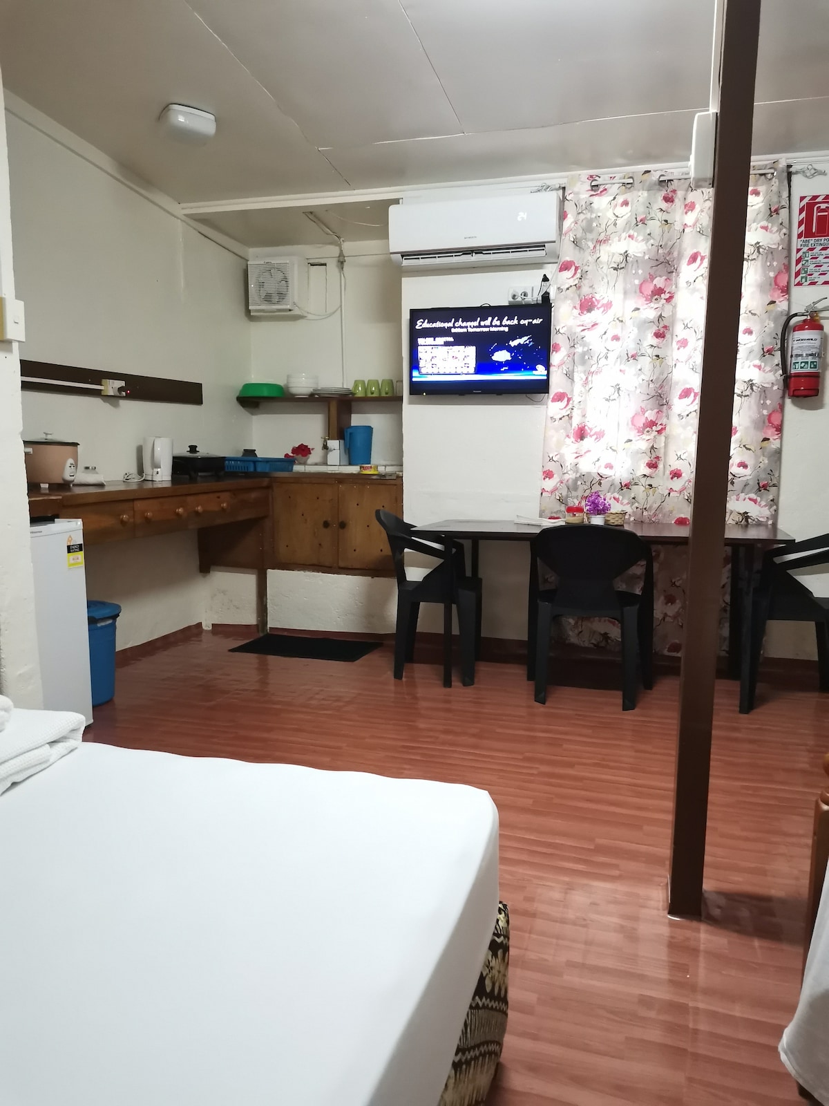 An airy room displays a bed with neatly arranged linens. A small dining area is visible, featuring two chairs and a table. A television is mounted on the wall, and a work area with wooden countertops and kitchen amenities is present in the background.