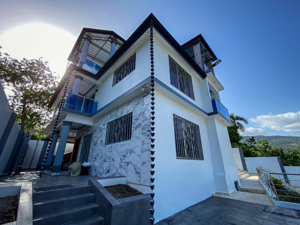 A three-story building features a clean, modern façade with a mix of white and stone accents. Large windows provide natural light, and the surrounding landscaping includes a paved entrance and a distant view of greenery.