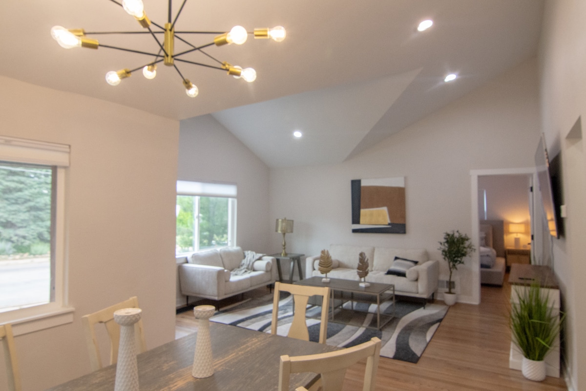 An open living area is featured with vaulted ceilings and modern decor. A dining table for six is positioned near the window, surrounded by stylish lighting. A cozy seating area is visible with two sofas and a central coffee table, contributing to an inviting atmosphere.
