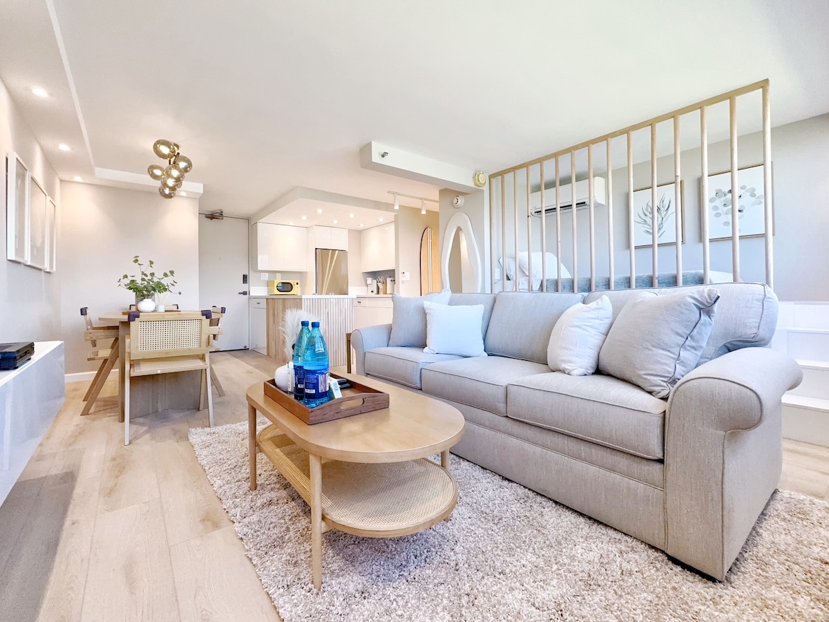 The living area showcases a light-colored sofa with soft pillows, placed in front of a round coffee table. A dining table is visible, accompanied by light wooden chairs. The background features the kitchen area with modern appliances, contributing to a relaxed, contemporary atmosphere.