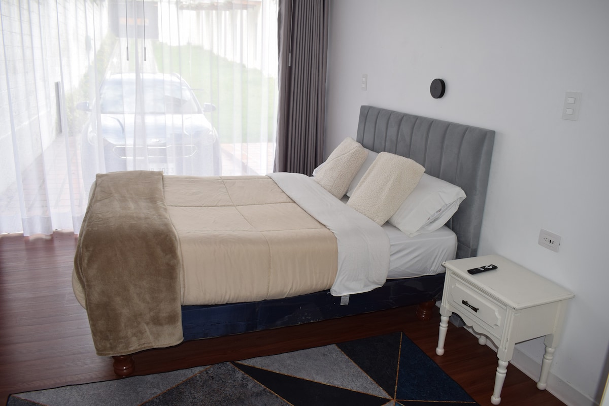 A comfortable sleeping area is furnished with a spacious bed topped with neutral-colored bedding. A bedside table is positioned beside the bed, featuring a remote control. Large windows with sheer curtains allow natural light to fill the room while providing a view of the outdoor space.