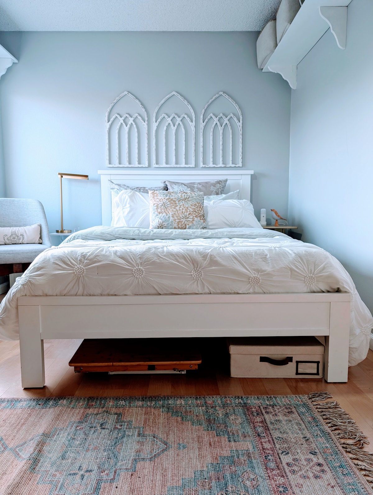 A cozy guest room features a spacious bed adorned with a decorative white quilt and accent pillows. A soft rug lies beneath the bed, while delicate wall features add character. A small nightstand holds a lamp, creating a serene atmosphere.