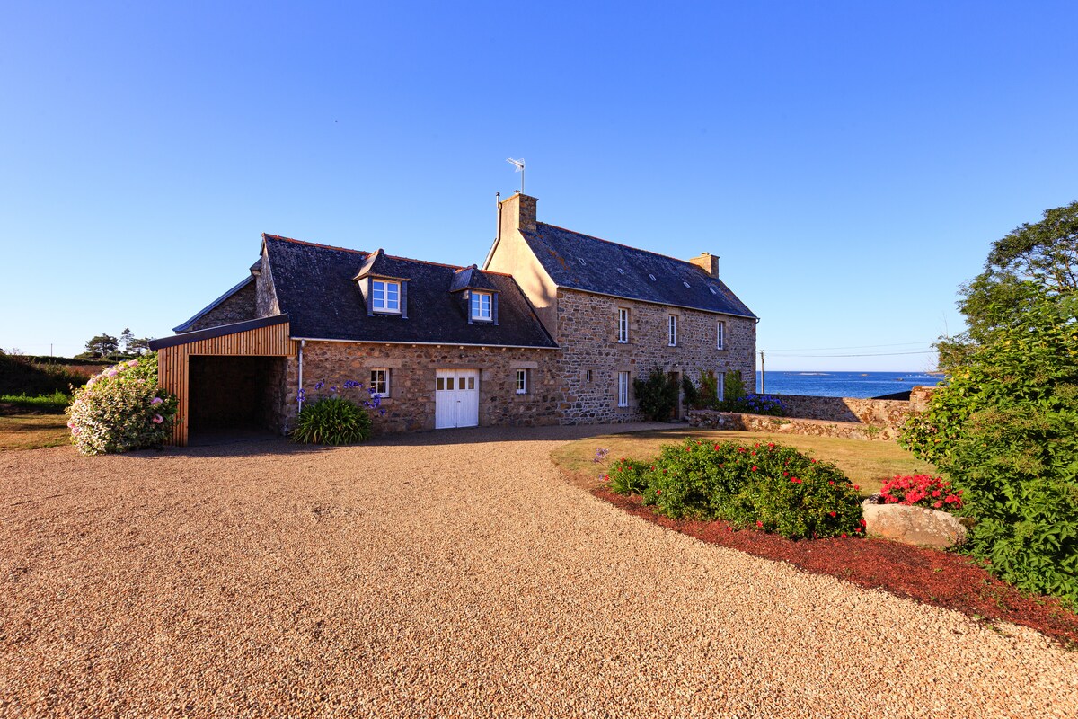 A charming stone house with a slate roof is presented against a clear blue sky. The property features a gravel driveway leading to a front entrance, with lush greenery and blooming flowers surrounding the grounds. The ocean is visible in the background, enhancing the serene landscape.