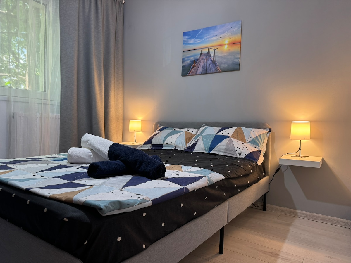 A modern bedroom features a spacious bed with a patterned quilt and neatly arranged towels. Soft light is provided by bedside lamps, enhancing the calming ambiance. A window allows filtered natural light, while a serene piece of wall art depicting a sunset adds a touch of color.