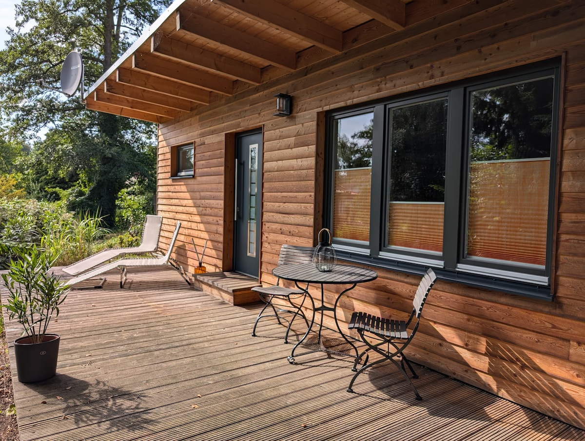 An inviting wooden terrace features a round dining table with black chairs and a relaxed lounge chair. Large windows provide views of the surrounding nature, while potted plants add a touch of greenery. The entrance door is framed by a modern light fixture.