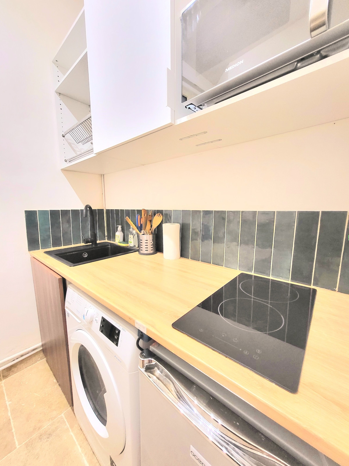 The kitchen area features a modern design with a wooden countertop. A washing machine is integrated into the cabinetry beside an electric cooktop. Above, shelves hold kitchen essentials, and a black sink is positioned next to cooking utensils, providing a functional and organized space.