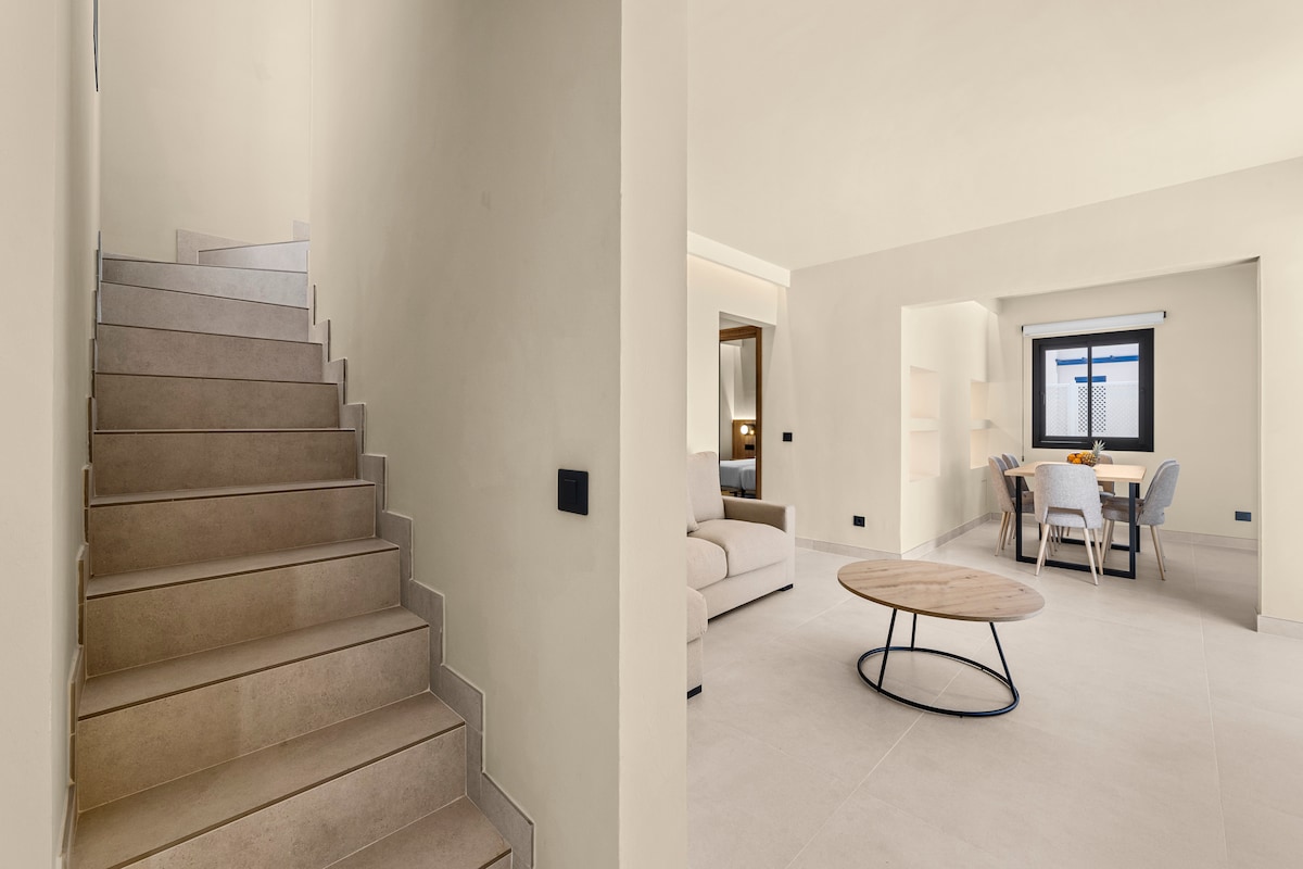 An open space is showcased, featuring a light-filled living area and dining setup. A staircase with neutral-colored steps leads to the upper levels. The dining table is surrounded by four chairs, and a soft-colored sofa rests in the living area.