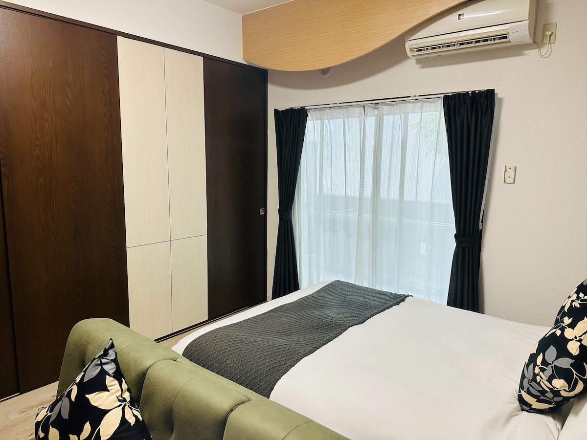 A comfortable sleeping area is presented with a spacious bed covered in crisp linens. Black curtains frame a large window, allowing natural light to brighten the room. A stylish wardrobe offers ample storage, complemented by decorative cushions on the green upholstered headboard.