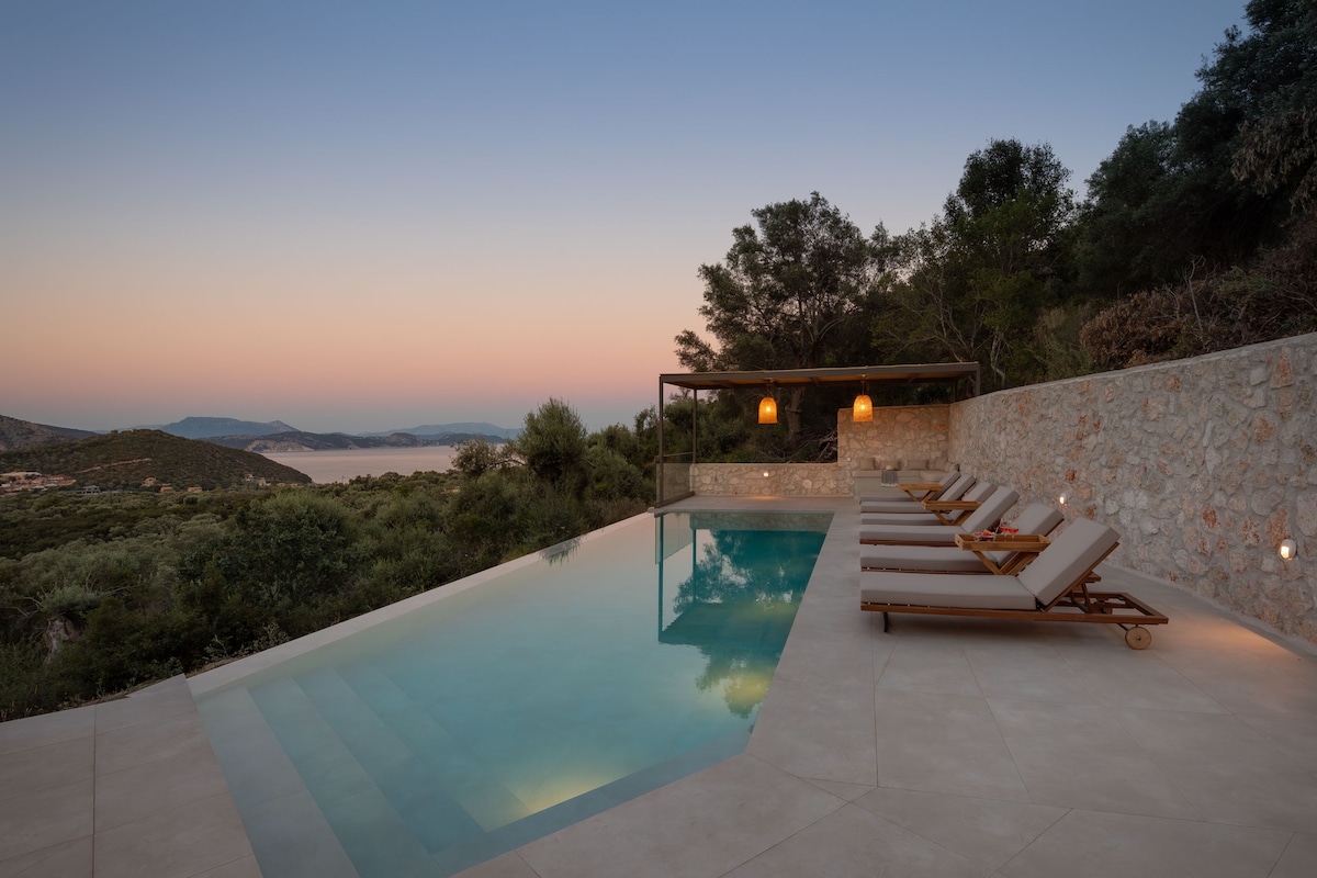 An inviting outdoor pool area features a uniquely shaped private pool lined with sun loungers. A bamboo pergola provides shade, while distant hills and water are visible in the background, creating a serene atmosphere at dusk.