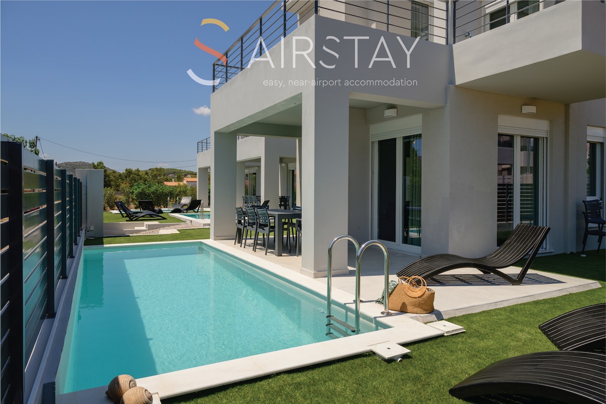 A private pool is surrounded by sun lounges and a grassy area, creating an inviting outdoor space. The modern maisonette is visible, with large glass doors offering views of the pool and shaded dining area. The clear blue sky enhances the tranquil atmosphere.