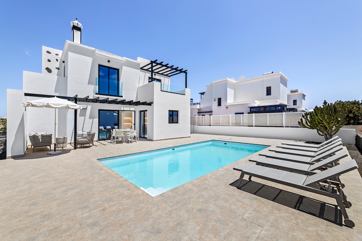 A modern villa is framed by a spacious outdoor area, featuring a heated swimming pool surrounded by multiple lounge chairs and shaded seating. A dining area is visible near the pool, complemented by a shaded umbrella and a view of surrounding structures.