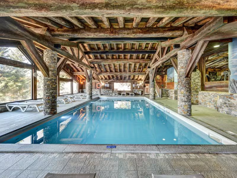 An indoor pool area is framed by wooden beams and stone pillars. Lounge chairs are arranged along the edges, while large windows allow natural light to illuminate the water. The serene environment emphasizes relaxation and wellness within the residence.