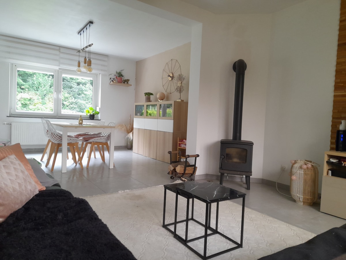 A bright living area features a stylish dining table with chairs, complemented by a cozy seating arrangement. A modern stove is positioned near the wall, and large windows allow natural light to illuminate the space, offering views of the surrounding greenery.