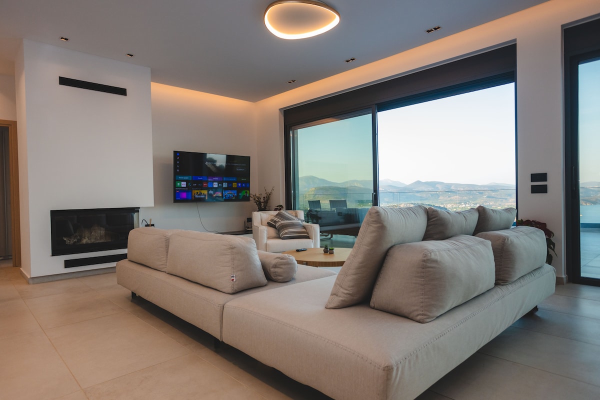 A spacious living area is furnished with a large, soft sectional sofa featuring plush cushions. A wall-mounted television is visible along with a minimalistic fireplace. Floor-to-ceiling windows showcase serene views of the landscape and mountains beyond, allowing ample natural light to fill the room.