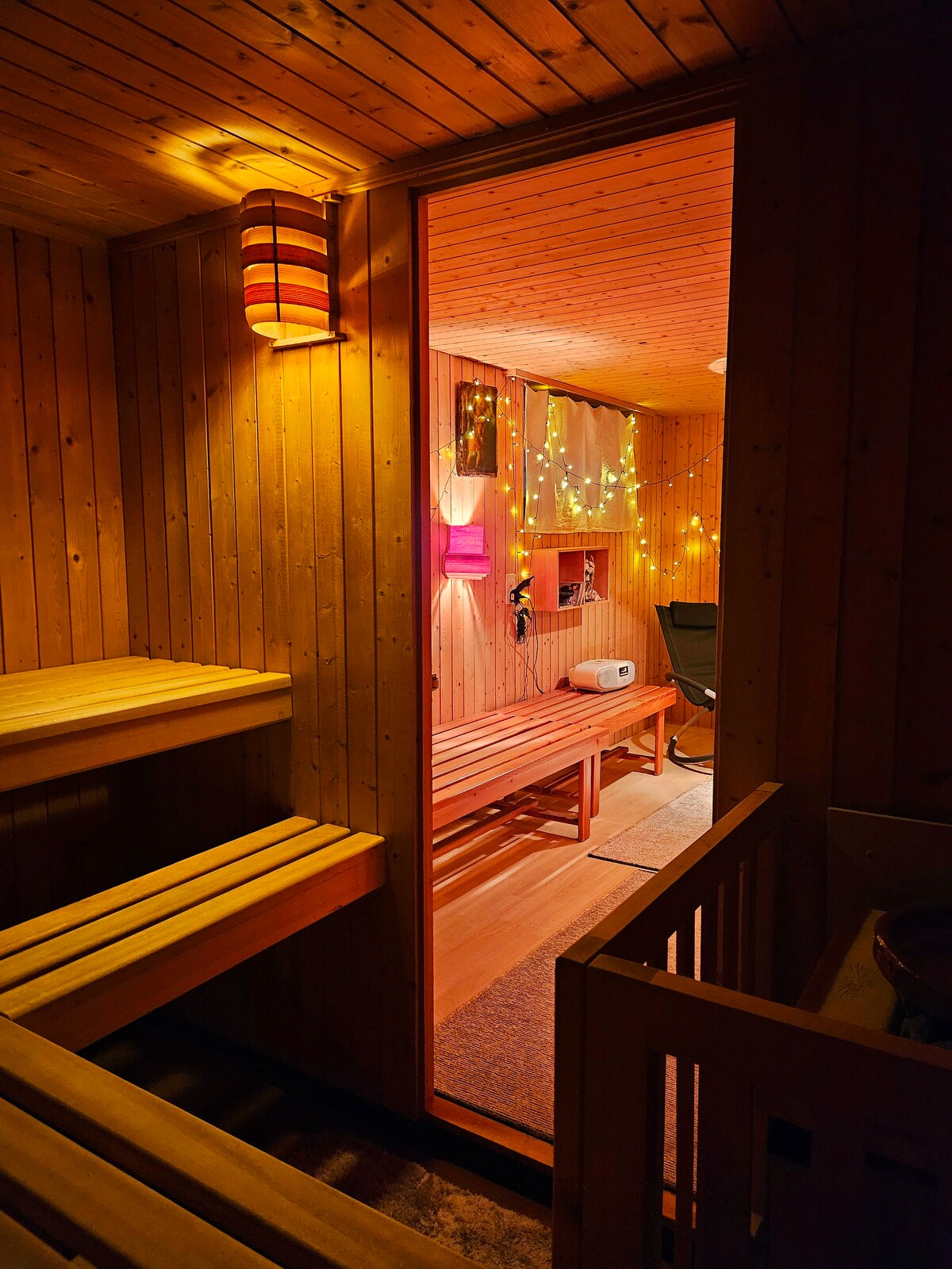 A wooden sauna space features benches for seating, illuminated by soft ambient lighting. An open doorway leads to a cozy relaxation area decorated with fairy lights and a chair. The warm wood tones enhance the inviting and calming environment.